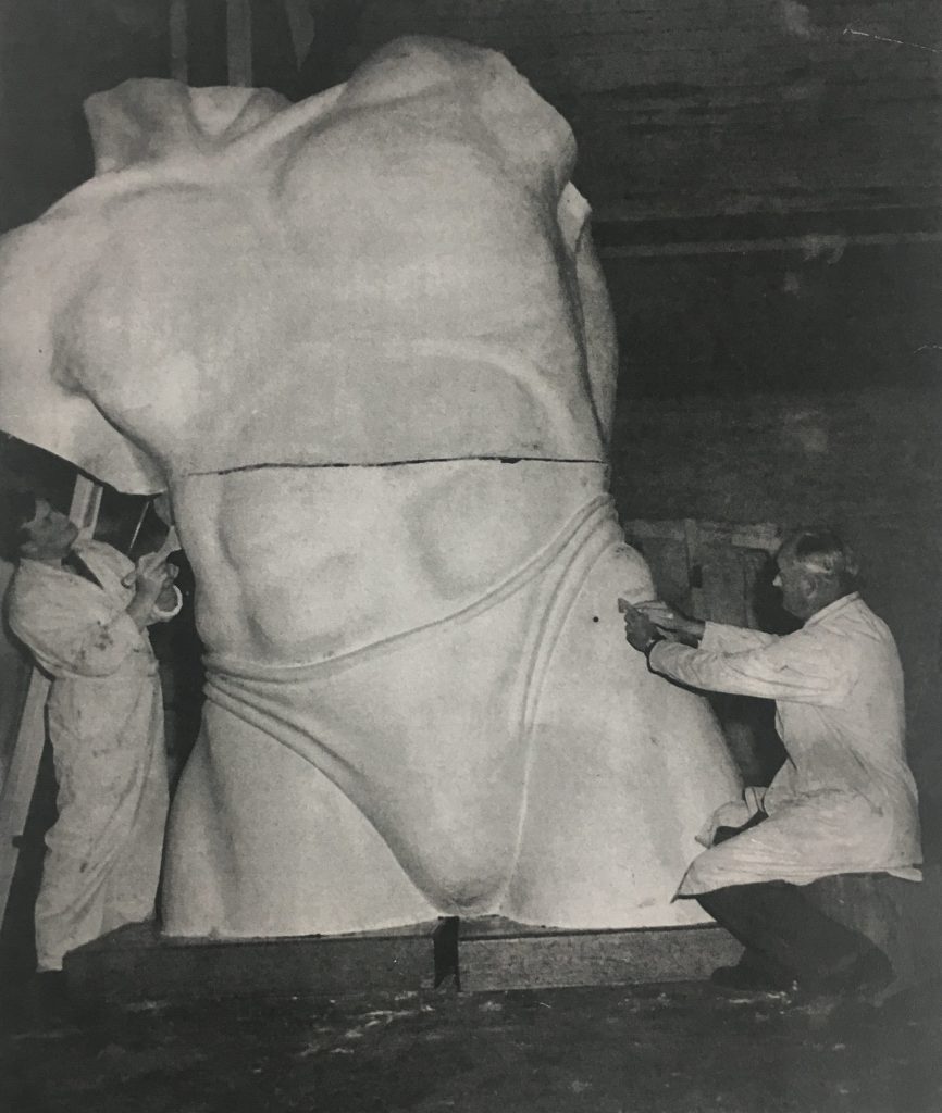 An image of a large torso made of plaster, split into two pieces. Two craftsman work on the torso, which is twice their height.