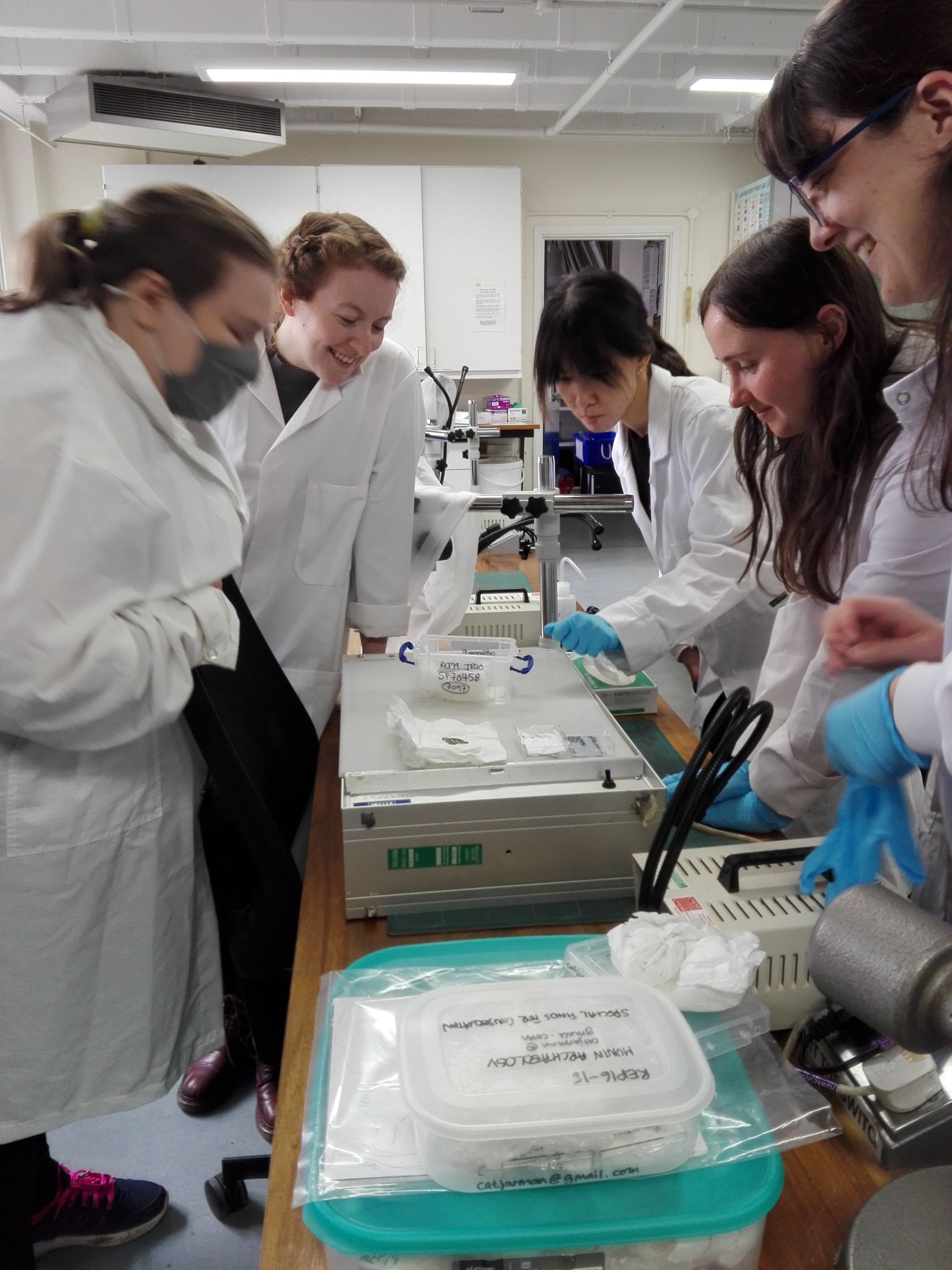 Five students in white lab coats gathered around a small open plastic box on a table, peering into it with smiles on their faces.