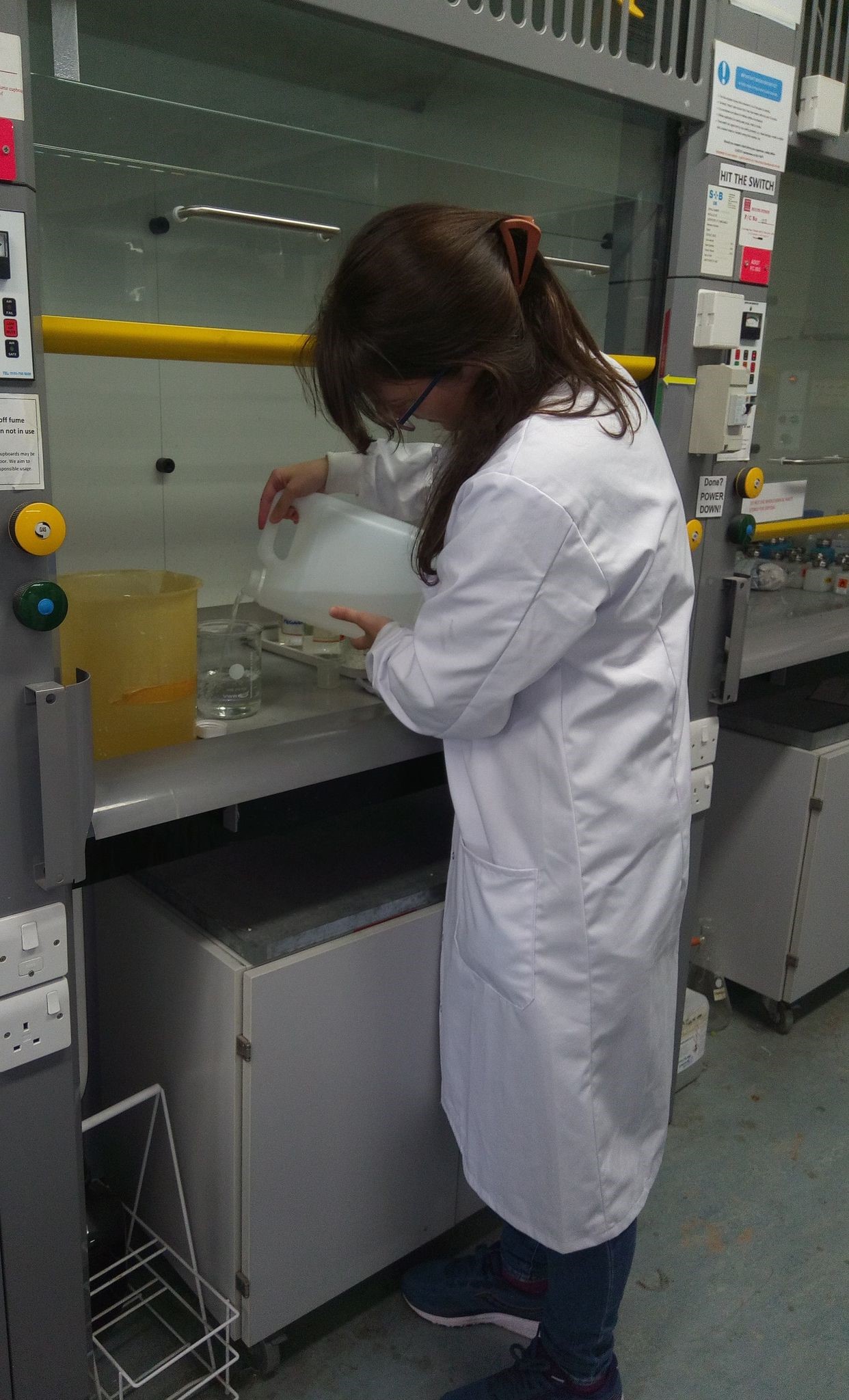 A woman with long brown hair in a white coat pours a liquid into a glass container standing on an electric balance inside a fume cupboard.