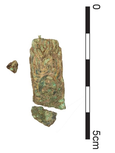 A pale gold-coloured rectangular strap and two fragments decorated with a knotwork pattern. There are green and blue patches of corrosion between the raised sections of the knotwork. The scale shows that the strap is just over 3cm long in total, including fragments, and just under 2cm wide.