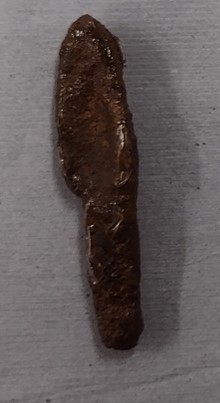 A long elliptical object with a hollow in the middle and a small rectangular handle, resembling a scoop. The object is covered in dark brown corrosion. 