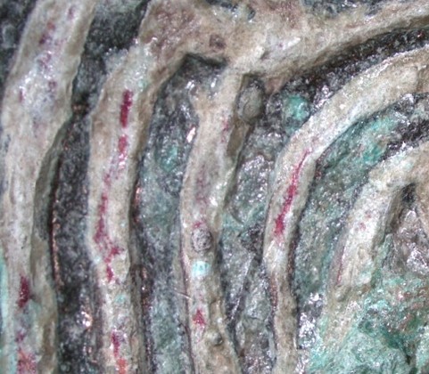 A close up of several dark red streak-shaped deposits between the raised sections of knotwork. 
