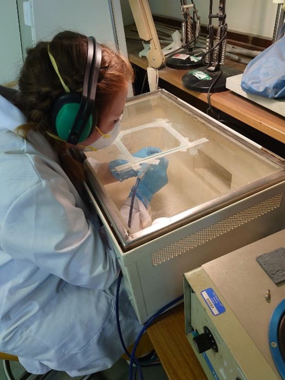 A student with red hair in a plaited bun wearing a white lab coat, ear defenders and a face mask sits in front of an air abrasion cabinet. She looks through the glass panel on the upper face of the cabinet at the abrasive nozzle she is holding in gloved hands as she guides it over the surface of an iron object. 