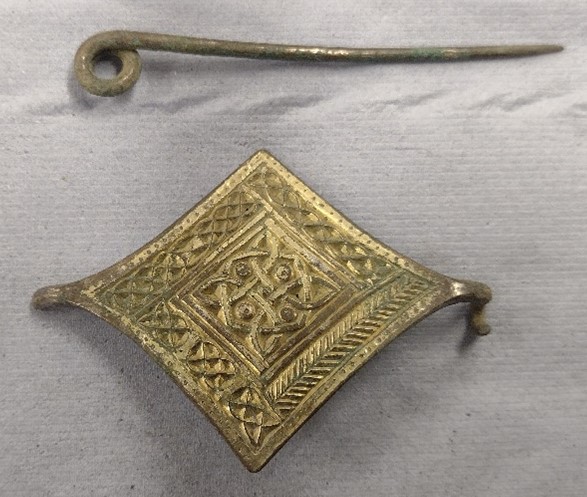 A gold-coloured diamond-shaped brooch with a central diamond shaped knotwork motif surrounded by a border of four different styles of knotwork, one along each side. Above, there is a long, dull coloured pin, one end bent into a circle shape. 