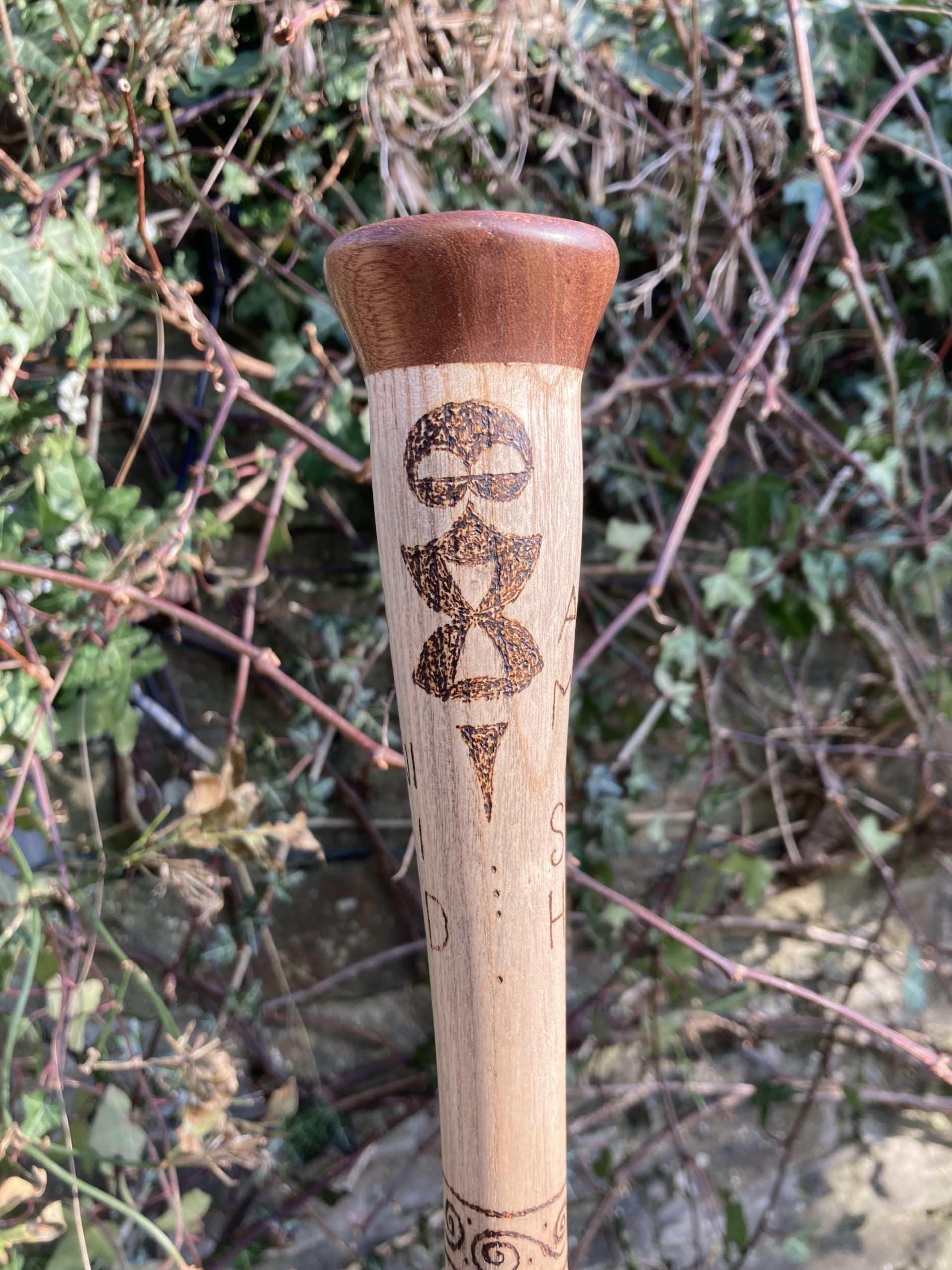 Dark pyrographic heavy lined images on a wooden staff, inspired by Iron Age Celtic art. Blurred background is plants and dirt.