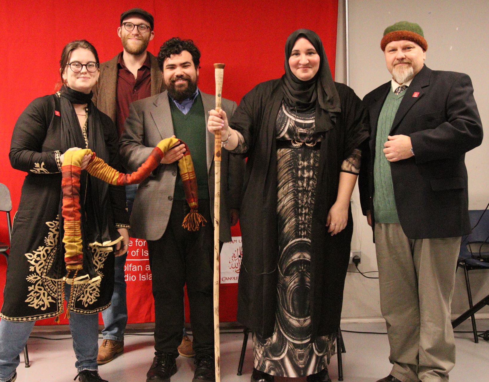 A group of people stand before a red banner. In the centre, a Muslim woman poet in hijab carries a crafted and decorated wooden staff. On either side are men wearing wool jumpers, blazers, and beards. The younger one is next to a young woman in glasses, and they carry between them a fabric bag. Behind them, a taller bespectacled man with beard and cap is standing. All are smiling.