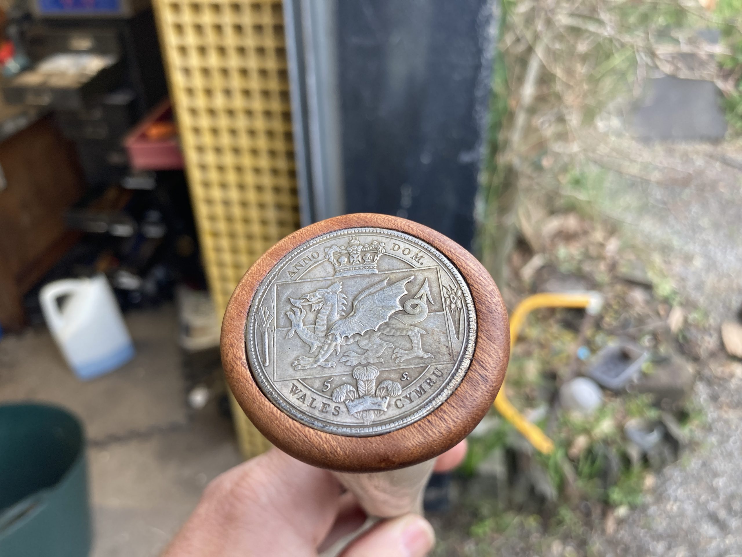 A metal coin embossed with a dragon is seen embedded into the top of the wooden staff. It sits just below centre, with the hand holding the staff visible. The background is blurred with use of aperture but you see the garage wall in the centre with garage on the left and gravel to the right.