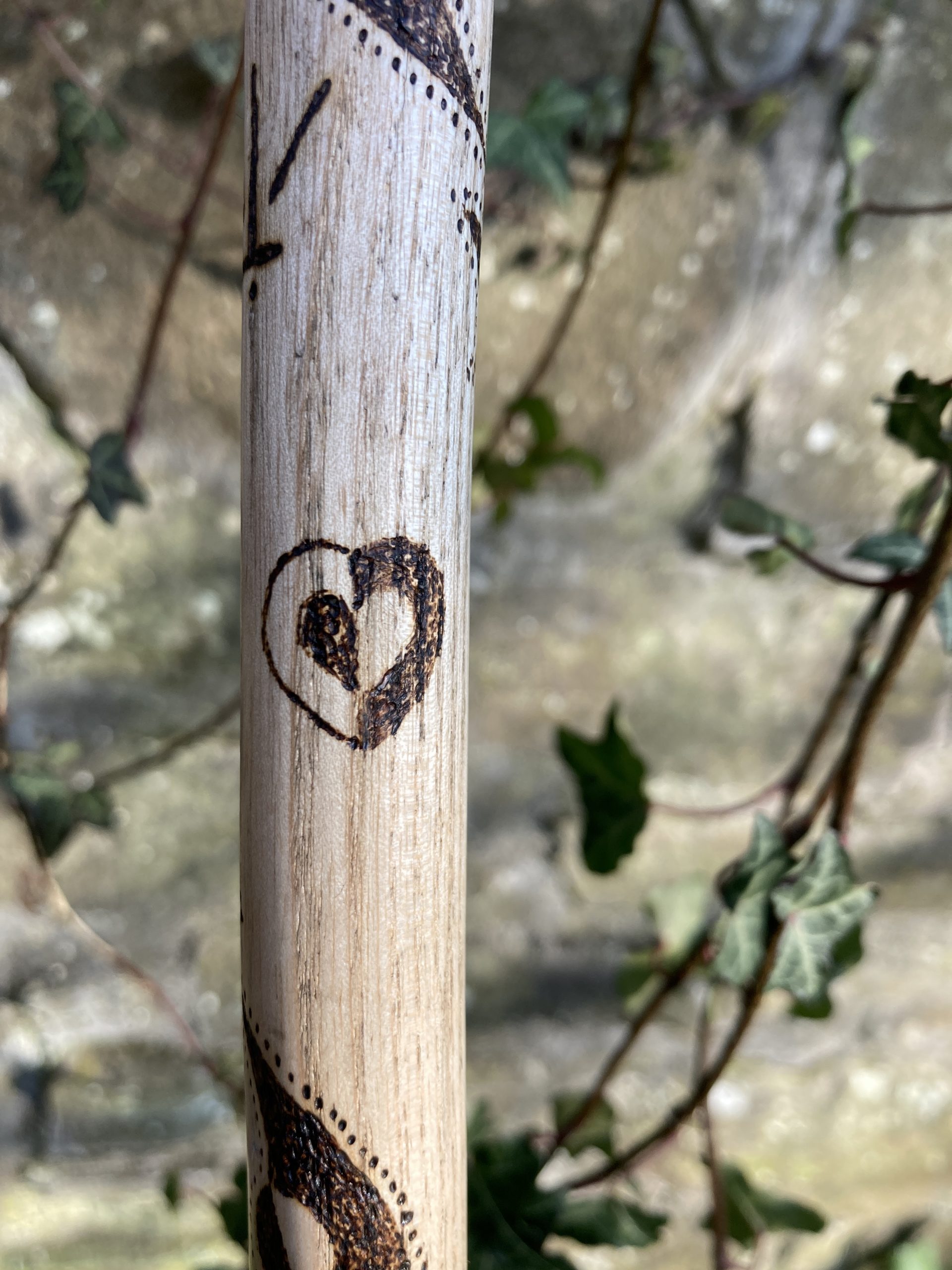 An image of a heart pyrographed onto the wooden staff. It plays with negative inversion, with a dark outline, wood-toned outer image, and dark centre on the left side of the heart. The right side is the opposite with a dark exterior and pale interior.