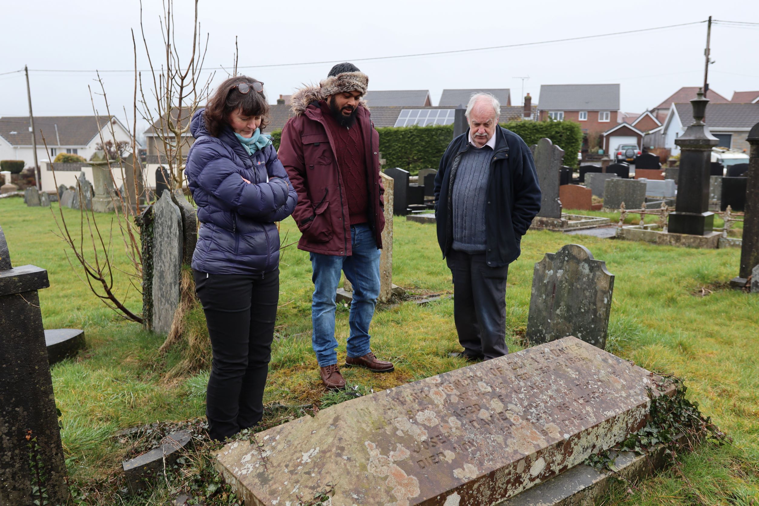 Three people in warm coats look at a gravestone in a church cemetery on a grey day in Wales. A woman on left clutches books in her arms; a young man in centre wears a red coat and furry warm hat; an older man on the right has his dark jacked open and his hands in his pockets. 