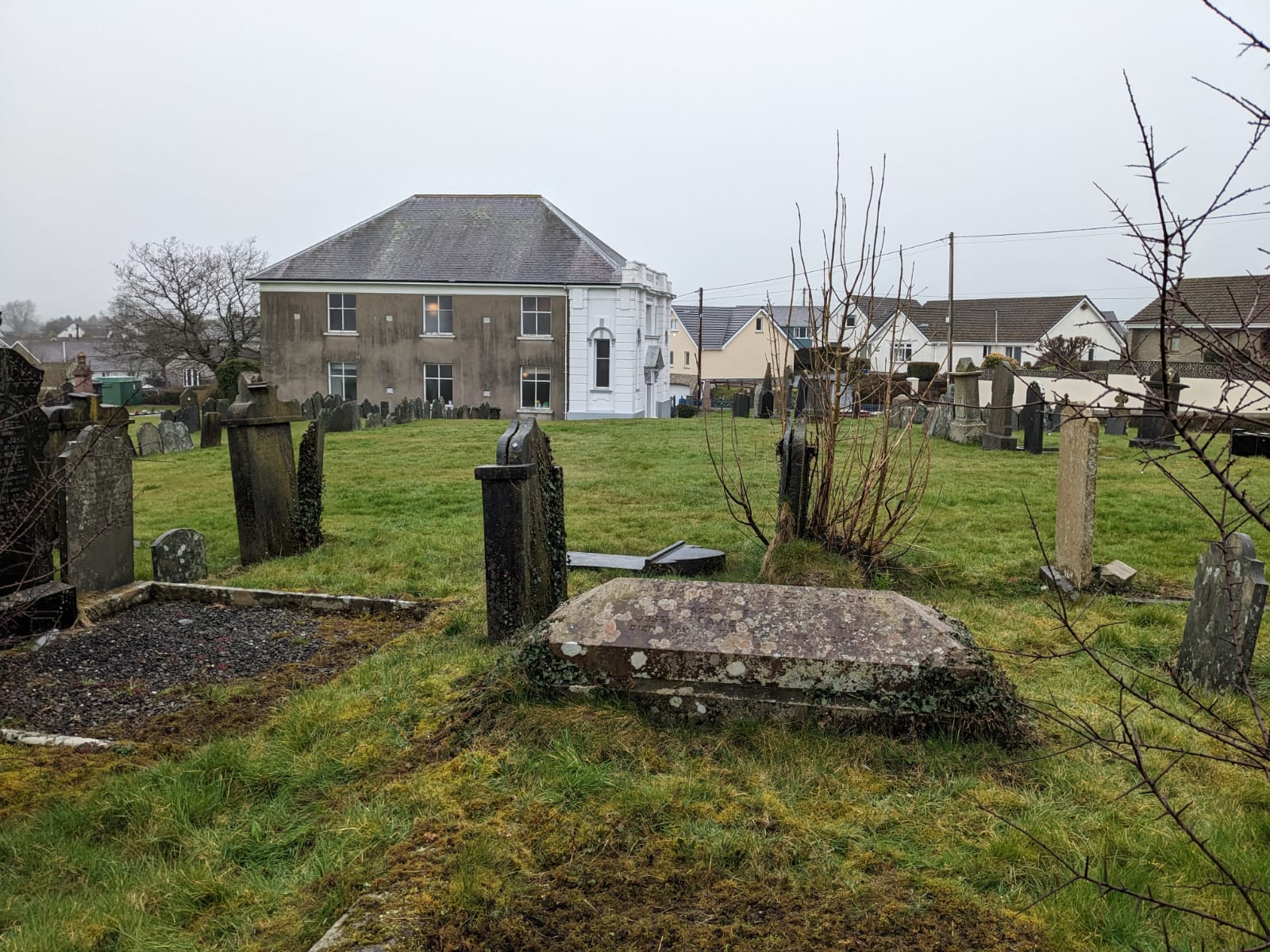 The chapel of Bethlehem St Clears in Carmarthenshire - a white front on a concrete building with six windows over two storeys showing in the middle distance. Several graves are in the foreground, with headstones and one large grave cover, and a green field is in the photo's centre.