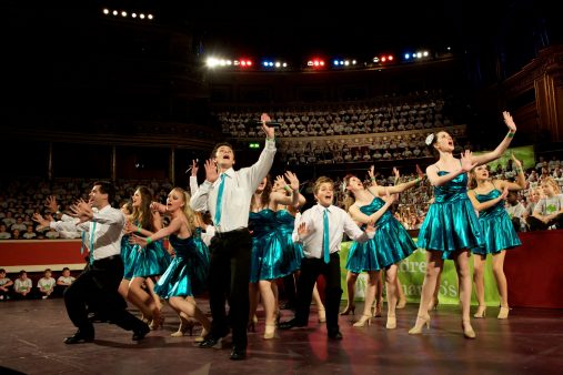 A group of men, women, boys and girls singing on a stage in front of a large audience.