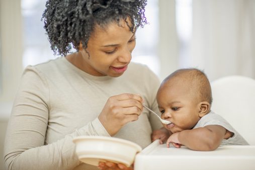 A young mother is spoon feeding her son in the kitchen.