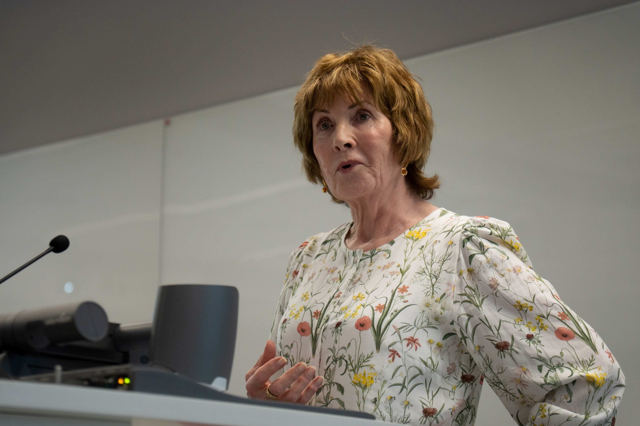 A woman with short hair and wearing a flowery top, standing at the lectern