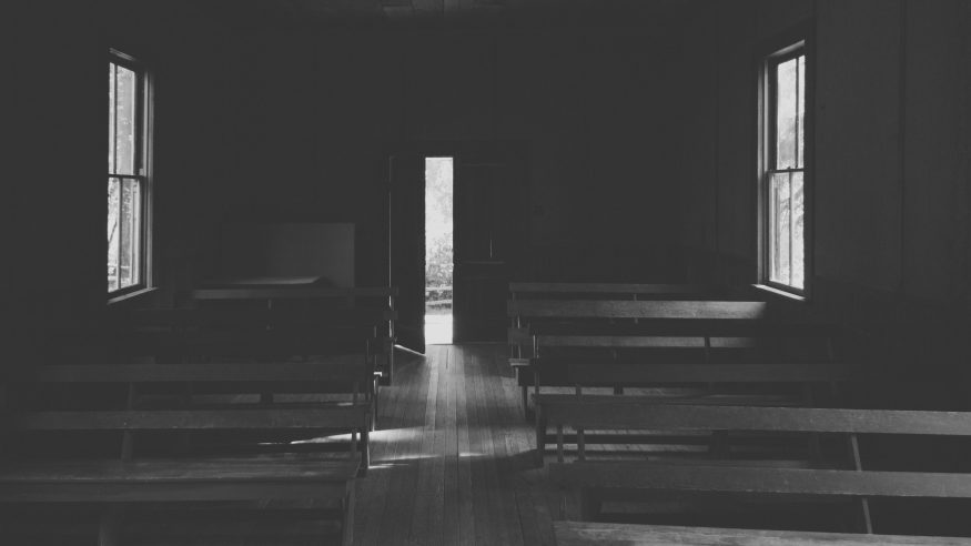 An interior of a small church in the countryside with wooden benches and an opened door shot in black and white