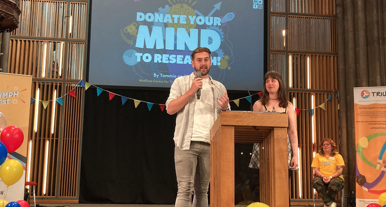 A young man and woman on stage in front of a large screen which reads Donate your mind to research